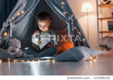 Using flashlight to read the book. Cute little girl playing in the tent that is in the domestic room 123006203