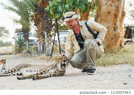 Male tourist in casual clothes and hat sitting and petting a cheetah outdoors Male tourist in casual clothes and hat sitting and petting a cheetah outdoors 123007463