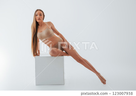 Sitting on the one leg on the white cube. Woman in underwear with slim body type is posing in the studio Sitting on the one leg on the white cube. Woman in underwear with slim body type is posing in the studio 123007938