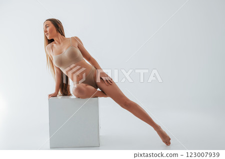 Sitting on the one leg on the white cube. Woman in underwear with slim body type is posing in the studio 123007939