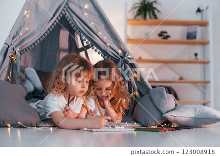 Learning to write and draw. With paper and pencils. Two little girls is in the tent in domestic room together 123008918
