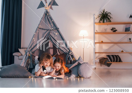 Laying down and using flashlight to read the book. Two little girls is in the tent in domestic room together Laying down and using flashlight to read the book. Two little girls is in the tent in domestic room together 123008930