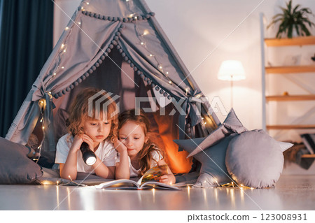 Laying down and using flashlight to read the book. Two little girls is in the tent in domestic room together 123008931