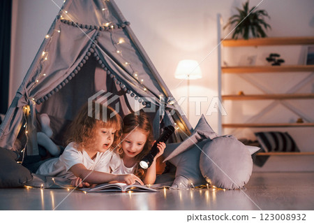 Laying down and using flashlight to read the book. Two little girls is in the tent in domestic room together 123008932