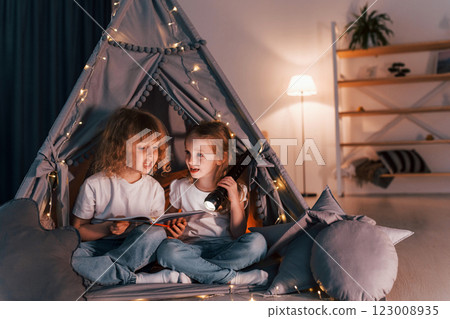 One girl holding flashlight, other reading the book. Two little friends is in the tent in domestic room together One girl holding flashlight, other reading the book. Two little friends is in the tent in domestic room together 123008935