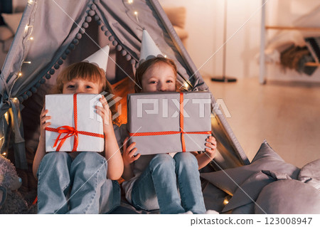 Holding gift boxes. Conception of presents and holidays. Two little girls is in the tent in domestic room together 123008947