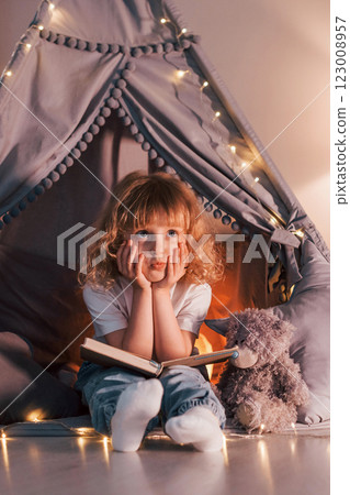Sitting with book. Cute little girl with curly hair is in the tent in domestic room Sitting with book. Cute little girl with curly hair is in the tent in domestic room 123008957