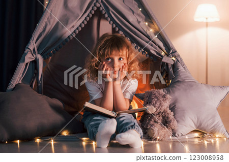 Sitting with book. Cute little girl with curly hair is in the tent in domestic room Sitting with book. Cute little girl with curly hair is in the tent in domestic room 123008958