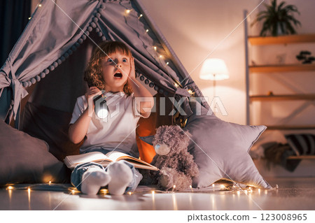 Wondered by book. Using flashlight. Cute little girl is in the tent in domestic room 123008965
