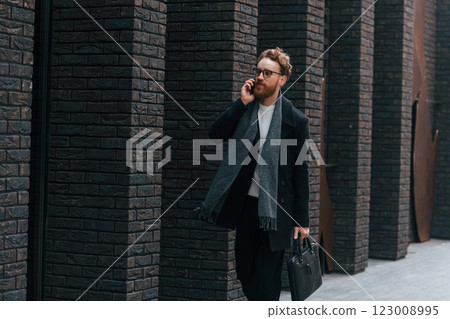 Talking by phone. Stylish man with beard and in glasses is outdoors near building 123008995