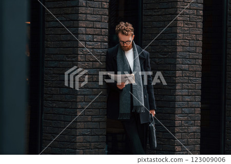 With newspaper in hands. Stylish man with beard and in glasses is outdoors near building With newspaper in hands. Stylish man with beard and in glasses is outdoors near building 123009006