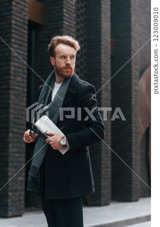 Holding newspaper in the hand. Stylish man with beard is outdoors near building Holding newspaper in the hand. Stylish man with beard is outdoors near building 123009010