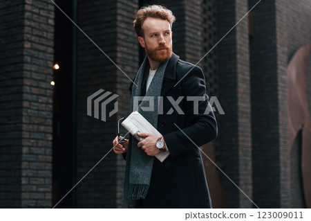 Holding newspaper in the hand. Stylish man with beard is outdoors near building 123009011