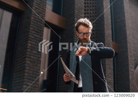 Checking time. Stylish man with beard and in glasses is outdoors near building 123009020