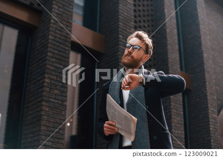 Checking time. Stylish man with beard and in glasses is outdoors near building 123009021
