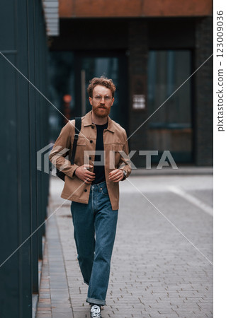 Front view. Stylish man with beard in khaki colored jacket and in jeans is outdoors near building Front view. Stylish man with beard in khaki colored jacket and in jeans is outdoors near building 123009036