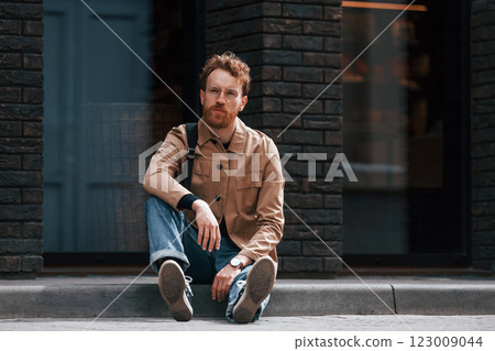 Sitting on the sidewalk. Stylish man with beard in khaki colored jacket and in jeans is outdoors near building 123009044