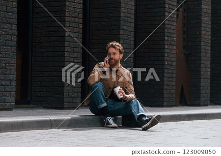 With phone. Stylish man with beard in khaki colored jacket and in jeans is outdoors near building 123009050