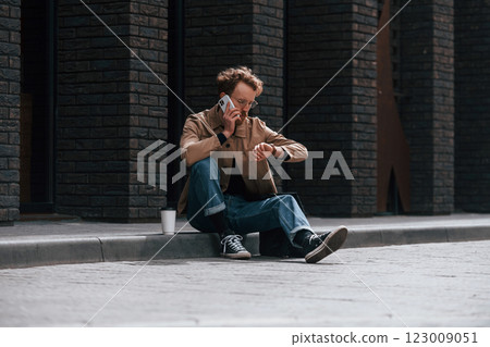 With phone. Stylish man with beard in khaki colored jacket and in jeans is outdoors near building 123009051