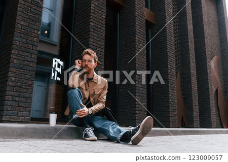 Coffee cup. Stylish man with beard in khaki colored jacket and in jeans is outdoors near building Coffee cup. Stylish man with beard in khaki colored jacket and in jeans is outdoors near building 123009057