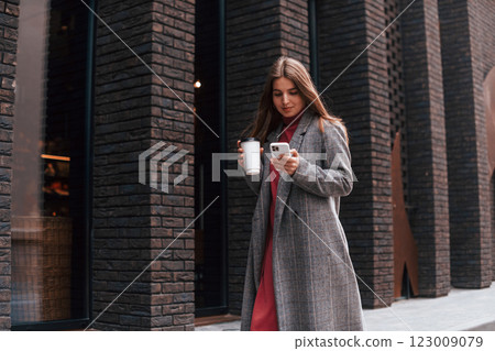 Young woman in grey colored coat is walking near building and holding smartphone and cup of drink Young woman in grey colored coat is walking near building and holding smartphone and cup of drink 123009079