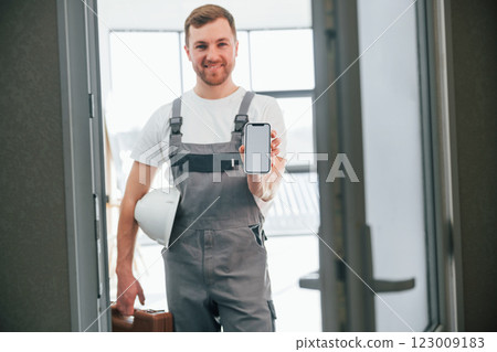 With smartphone in hand. Repairman is standing indoors in the modern room With smartphone in hand. Repairman is standing indoors in the modern room 123009183