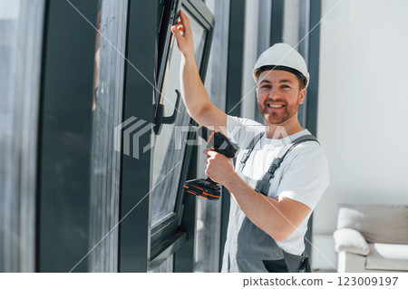 Using drill by installing windows. Repairman is working indoors in the modern room Using drill by installing windows. Repairman is working indoors in the modern room 123009197