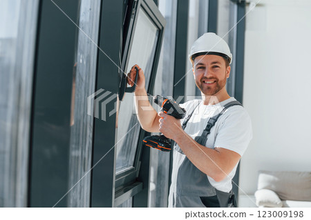 Using drill by installing windows. Repairman is working indoors in the modern room Using drill by installing windows. Repairman is working indoors in the modern room 123009198