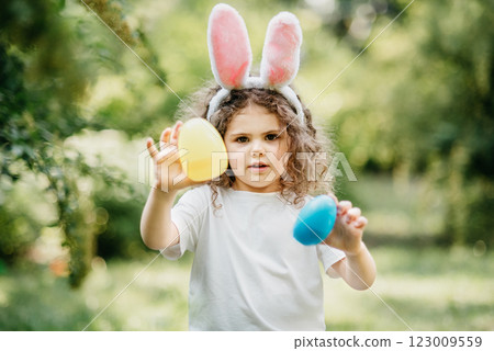 Easter egg hunt. Girl child Wearing Bunny Ears Running To Pick Up Egg In Garden. Easter tradition. Baby with basket full of colorful eggs. 123009559