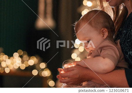 Mother with her little daughter are celebrating christmas indoors. Girl is holding garlands in hands 123010671