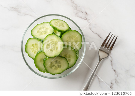 Fresh juicy cucumber slices in a glass bowl and metal fork over marble countertop. Fresh juicy cucumber slices in a glass bowl and metal fork over marble countertop. 123010705