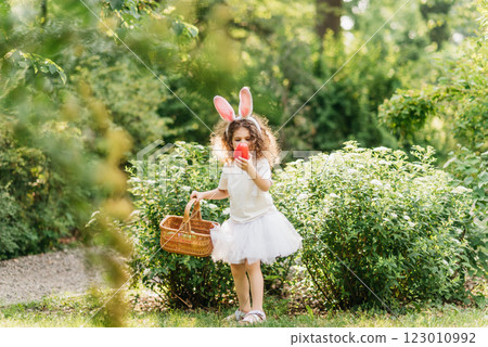 Easter egg hunt. Girl child Wearing Bunny Ears Running To Pick Up Egg In Garden. Easter tradition. Baby with basket full of colorful eggs. Easter egg hunt. Girl child Wearing Bunny Ears Running To Pick Up Egg In Garden. Easter tradition. Baby with basket full of colorful eggs. 123010992
