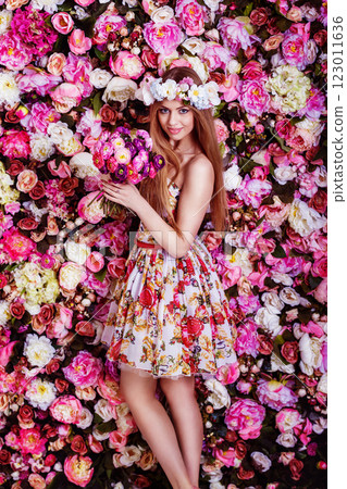 A beautiful young girl with flowers bouquet is standing near a floral wall. 123011636