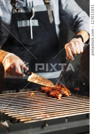 Chief cook is turning over meat at open fire grill at kitchen background. Vertical image. Chief cook is turning over meat at open fire grill at kitchen background. Vertical image. 123011831