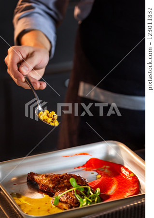 Chief cook is decorating a meat dish with corn at kitchen background. Vertical image. Chief cook is decorating a meat dish with corn at kitchen background. Vertical image. 123011832