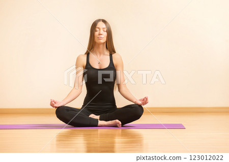 Young woman is practicing yoga and meditating sitting in lotus pose at gym background. 123012022