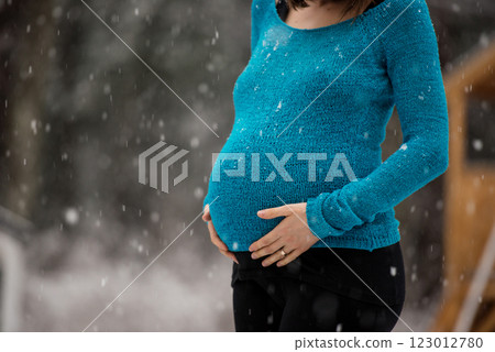 Pregnant woman in blue sweater standing outside on a snowy day touching her swollen belly 123012780