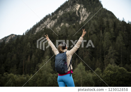 Young female hiker with a backpack standing under a mountain with her arms raised high in victorious gesture Young female hiker with a backpack standing under a mountain with her arms raised high in victorious gesture 123012858