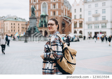 Tourist happy woman in glasses with backpack on Market Square in Krakow, Traveling Europe in autumn. St. Marys Basilica, cloudy day. Attractive female is exploring city. Top tourist attraction 123013504