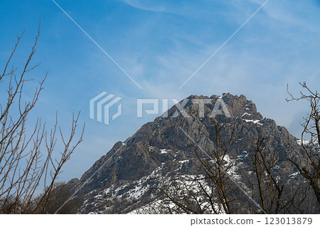 A natural mountaintop landscape with white small areas of snow-covered slopes against a blue sky with white smoky clouds. In the foreground there are winter branches of trees without leaves 123013879