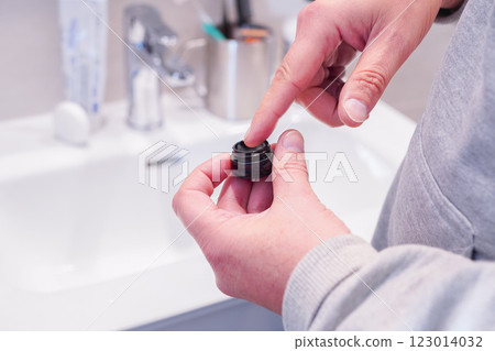 Close-up of a middle-aged man holding a container of beauty cream for skin care and applying cream to his face in front of a mirror 123014032