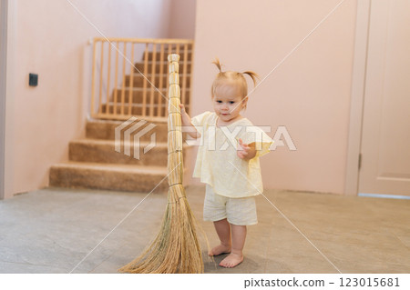 Portrait of barefoot toddler girl cleaning kitchen floor, gripping broom handle while assisting with housework in contemporary home setting, developing early sense of responsibility. 123015681