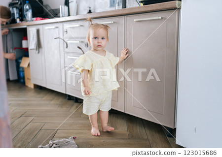 Full length portrait of curious toddler girl in yellow outfit exploring kitchen cabinets, touching them with hand while standing barefoot on wooden floor, looking at camera with happy expression. Full length portrait of curious toddler girl in yellow outfit exploring kitchen cabinets, touching them with hand while standing barefoot on wooden floor, looking at camera with happy expression. 123015686