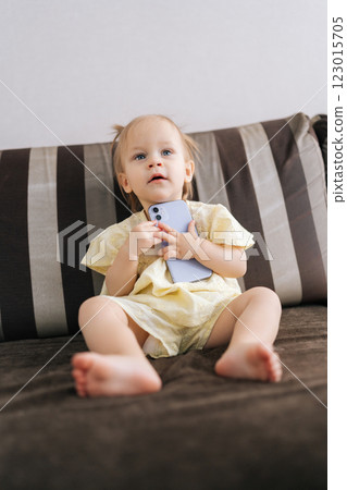 Vertical portrait of charming toddler girl sitting on comfortable sofa, holding smartphone and exploring digital world, demonstrating early age at which children interact with technology. Vertical portrait of charming toddler girl sitting on comfortable sofa, holding smartphone and exploring digital world, demonstrating early age at which children interact with technology. 123015705