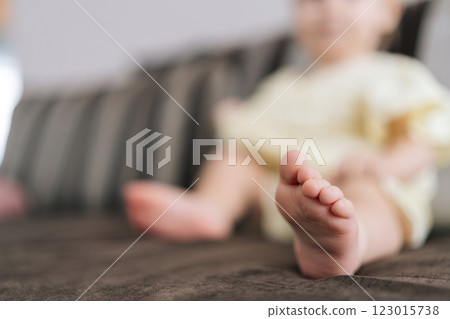 Close-up selective focus shot foot of baby girl wearing light yellow dress sitting comfortably on soft sofa in serene living room setting at home, cropped shot. Concept of curiosity and innocence. Close-up selective focus shot foot of baby girl wearing light yellow dress sitting comfortably on soft sofa in serene living room setting at home, cropped shot. Concept of curiosity and innocence. 123015738