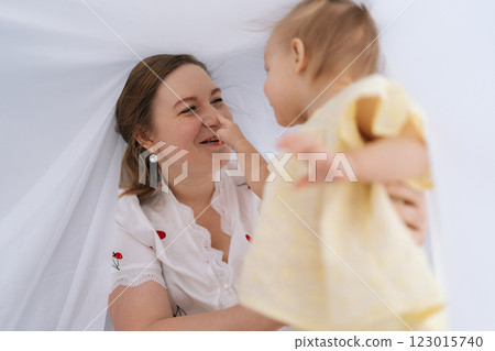 Cheerful young mother and infant daughter enjoying playful moments together, hiding under soft white blanket, pointing fingers and sharing laughter at home. Concept of parenting love and care. 123015740