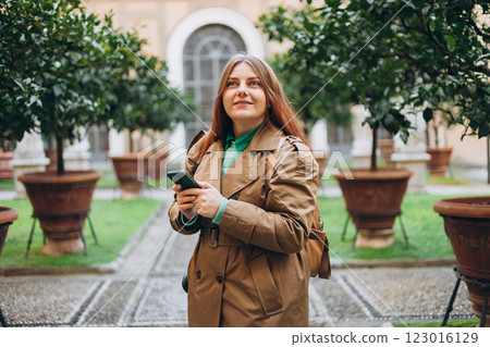 Happy 30s redhead woman using phone walking in city street in old town. Lifestyle, travel, tourism, technology, blogging, communication concept. Buildings with potted plants. Charming facade Happy 30s redhead woman using phone walking in city street in old town. Lifestyle, travel, tourism, technology, blogging, communication concept. Buildings with potted plants. Charming facade 123016129