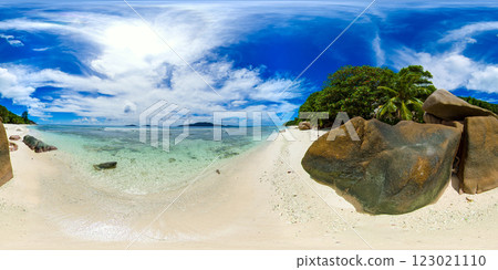 A serene beach with crystal-clear shallow waters, smooth boulders, and lush tropical vegetation, set against a vibrant blue sky. Anse Gaulettes. La Digue, Seychelles. VR 360. A serene beach with crystal-clear shallow waters, smooth boulders, and lush tropical vegetation, set against a vibrant blue sky. Anse Gaulettes. La Digue, Seychelles. VR 360. 123021110
