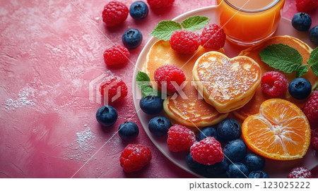 A romantic breakfast featuring fluffy pancakes topped with heart-shaped jam and surrounded by vibrant raspberries, blackberries, and blueberries, paired with a refreshing glass of orange juice. 123025222