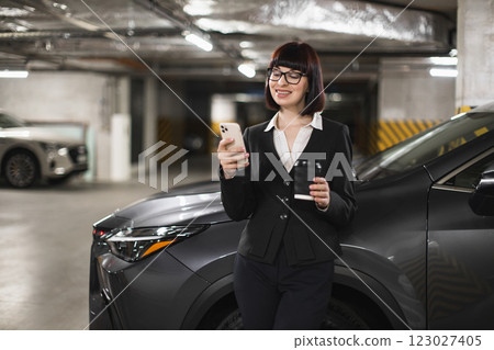 Young adult Caucasian female businesswoman in indoor car parking using smartphone with coffee in hand. Professional attire suggesting work or business meeting. Young adult Caucasian female businesswoman in indoor car parking using smartphone with coffee in hand. Professional attire suggesting work or business meeting. 123027405